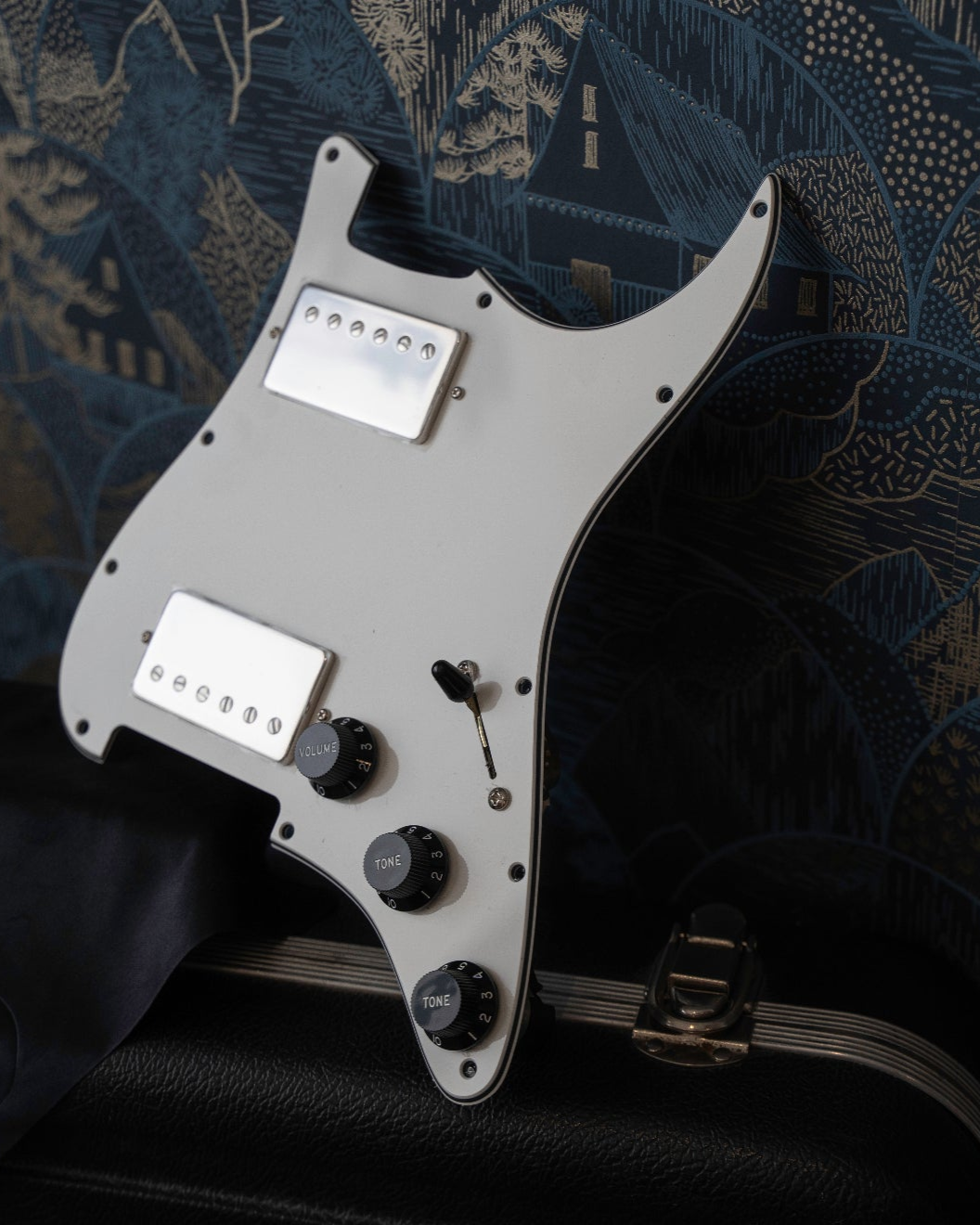 a parchment white loaded pickguard for HH Stratocaster is placed on top of a guitar case. The pickguard is rested against a wall with patterned blue and gold wall paper.