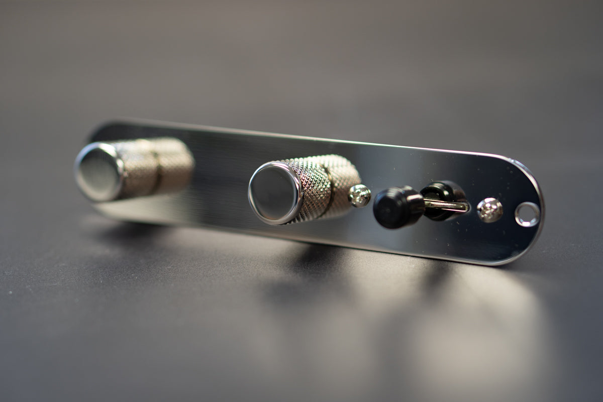 An artful view of an ObsidianWire telecaster loaded control plate with chrome knobs, chrome plate and a black switch tip. The control plate is set with a blurred grey background.