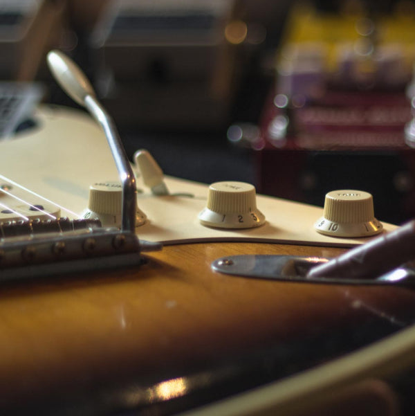 A close up shot of the switch, tone and volume controls on a Fender stratocaster. The image shows out of focus pedals in the back ground.