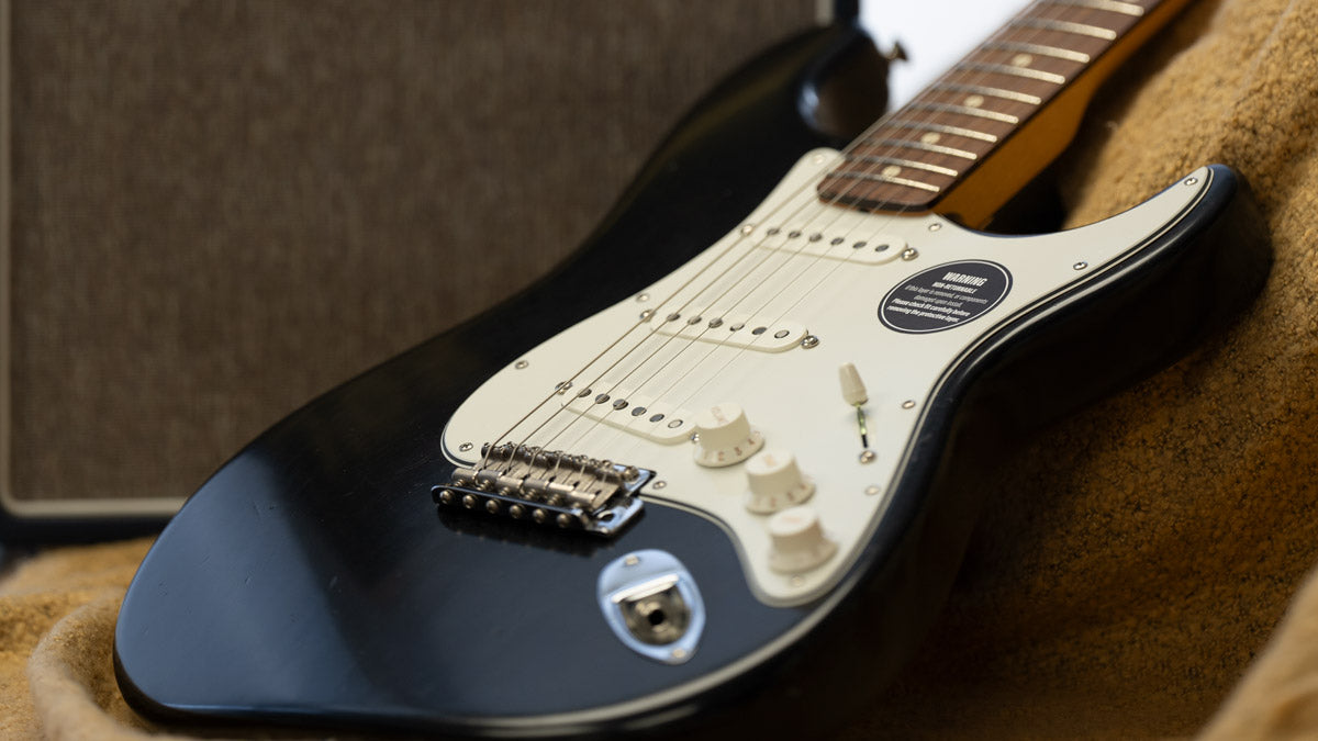 A black stratocaster with a parchment white loaded pickguard with parchment pickup covers and knobs. The stratocaster is set down on a blanket with amplifier grille cloth in the background.